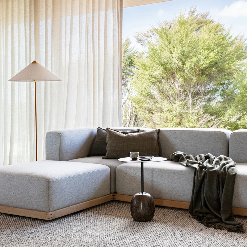 Modern living room with a light grey sectional sofa, slate grey linen cushion small table, and lamp near large windows.