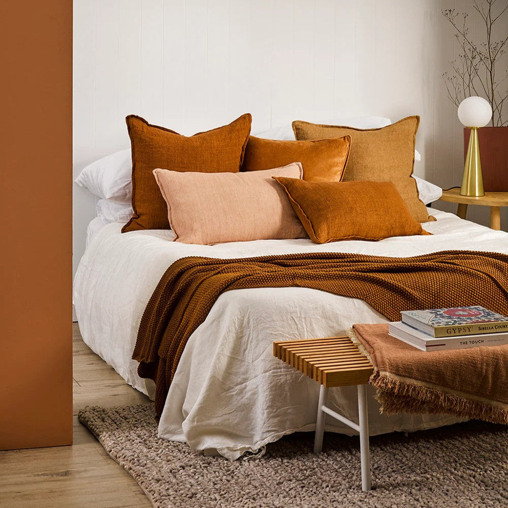 Bedroom with a bed featuring tobacco and salmon cushions and a blanket, next to a wooden side table.