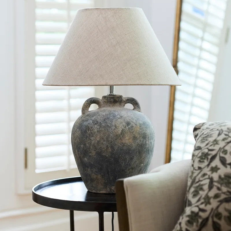 Table lamp with a textured base on a small round table in a room with light-coloured walls and a window with shutters.