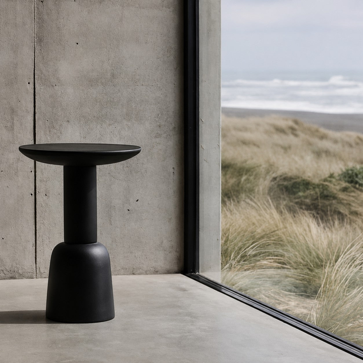 A matte black pedestal side table with a wide round top, positioned on a concrete floor next to a large window overlooking a beach dune landscape.