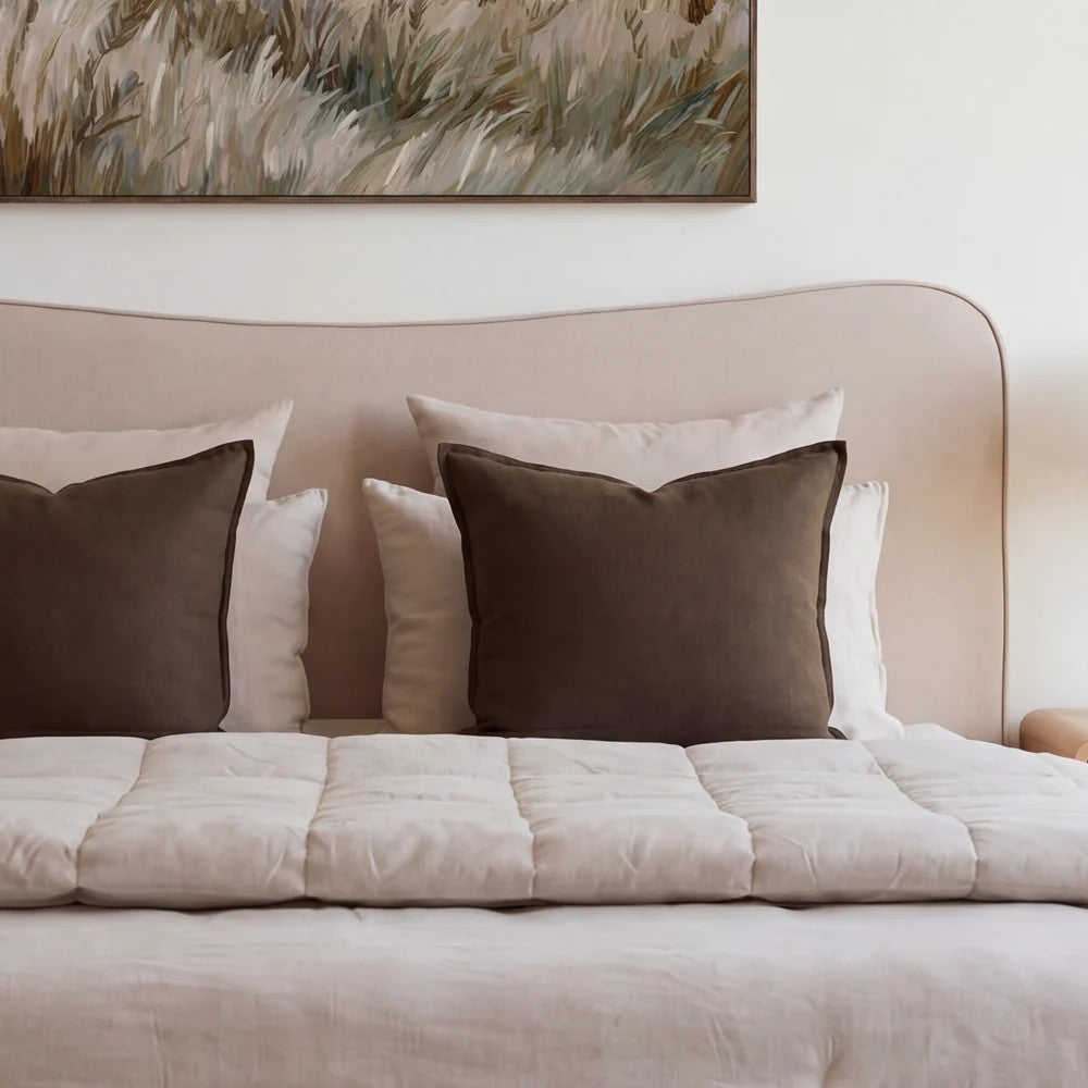 A styled bed with a beige upholstered headboard, cream bedding, and two dark brown accent pillows, with a framed abstract painting above on a white wall.
