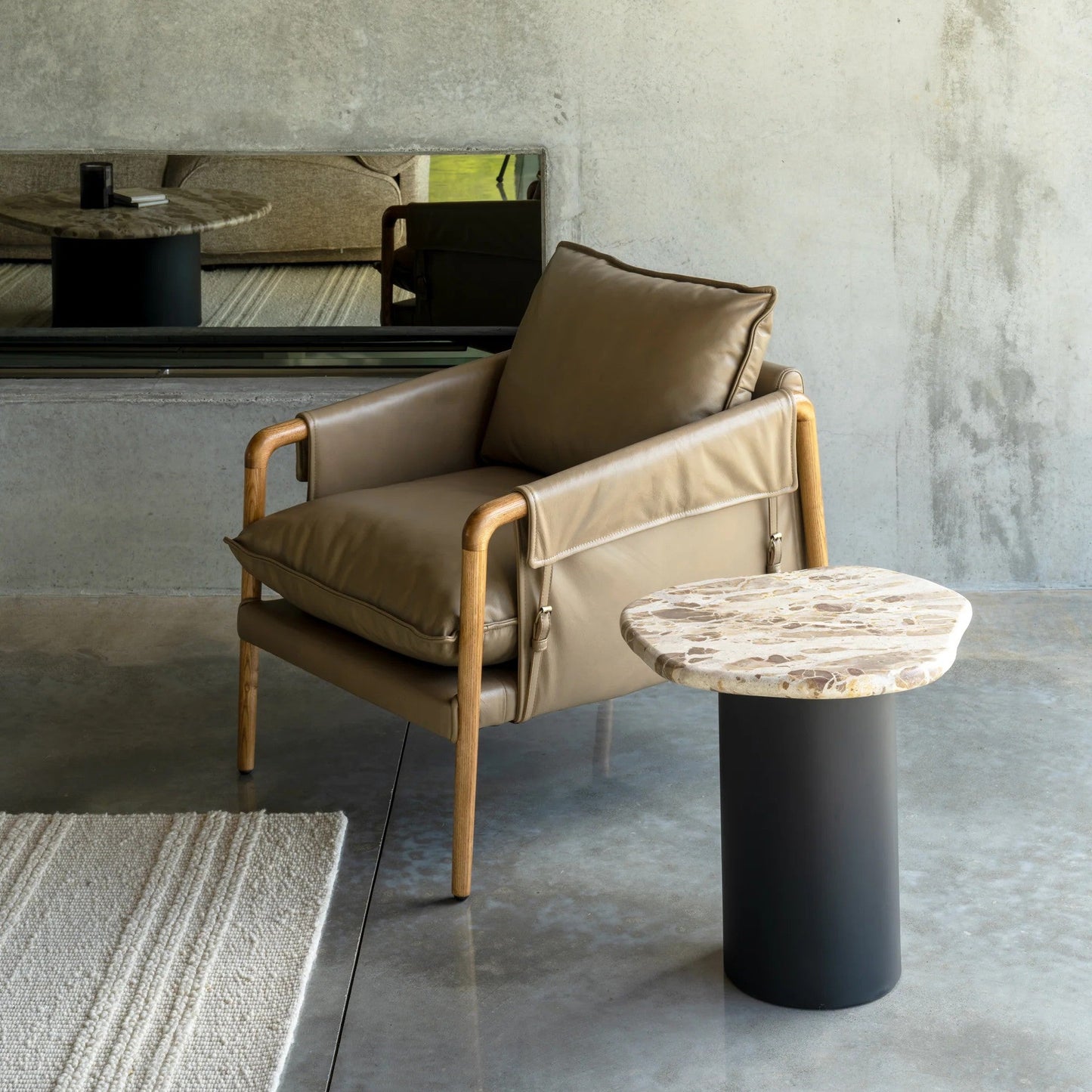 A modern tan leather armchair with light wood arms and legs, styled in a minimalist room with a textured concrete wall, a black fireplace, and a organic marble-top side table with a black base.