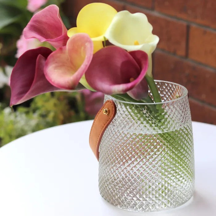 Clear textured vase with a faux brown leather handle holding pink, yellow, and red flowers against a blurred background.