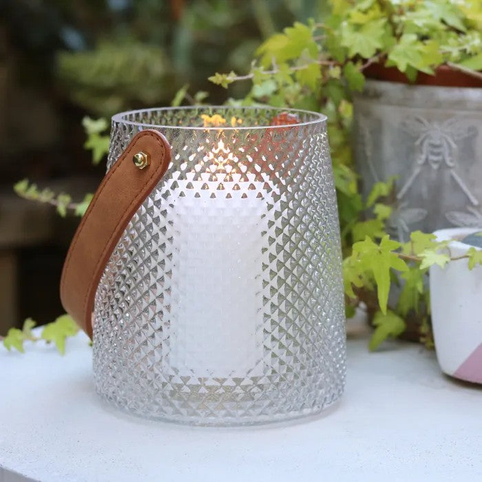 Decorative glass hurricane lantern with a brown faux leather strap on a table with plants in the background