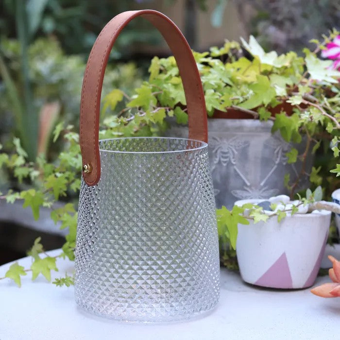 Glass Hurricane lamp with textured finish and brown faux leather handle n an outdoor setting