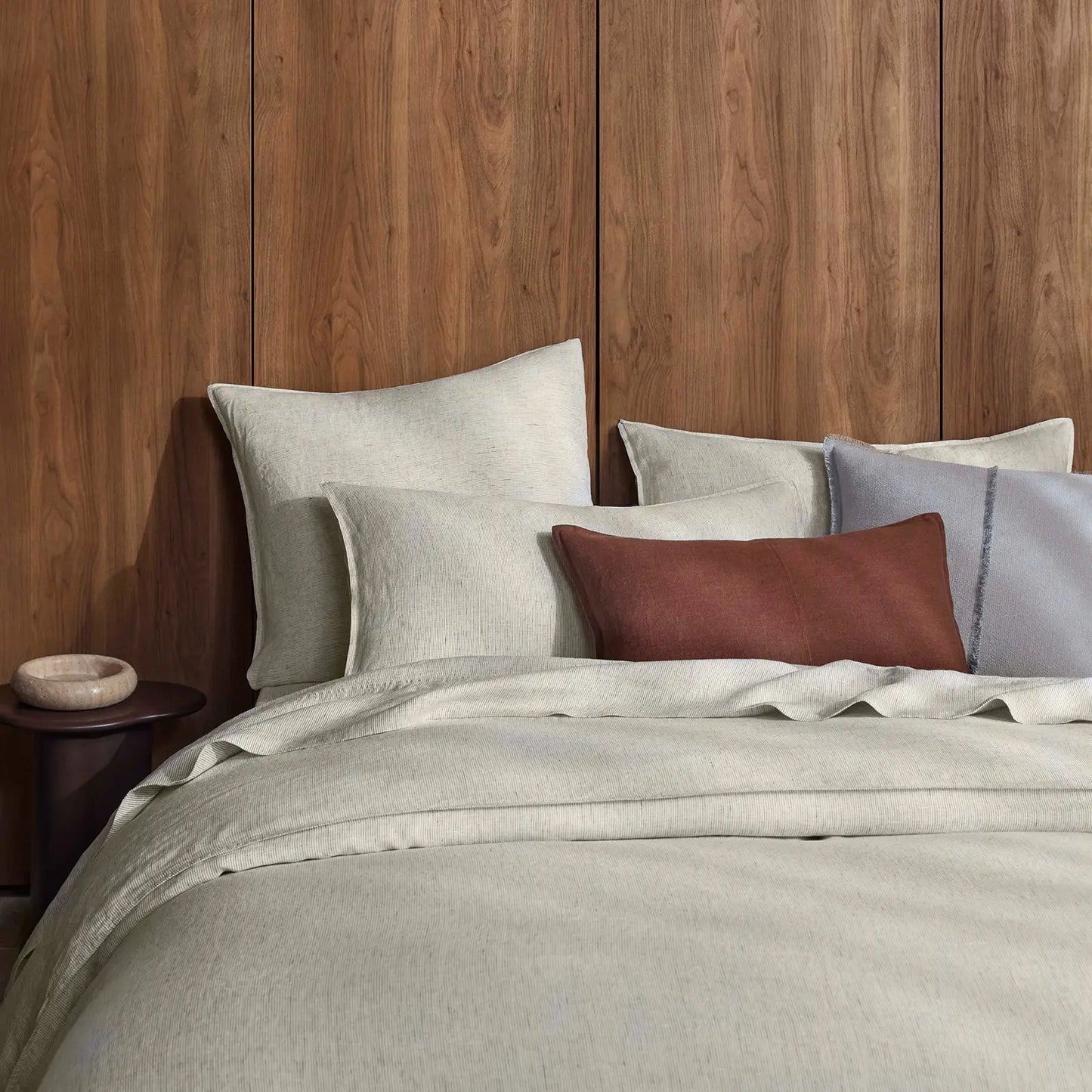 Neatly made bed with neutral bedding and cushions against a wooden panelled wall.