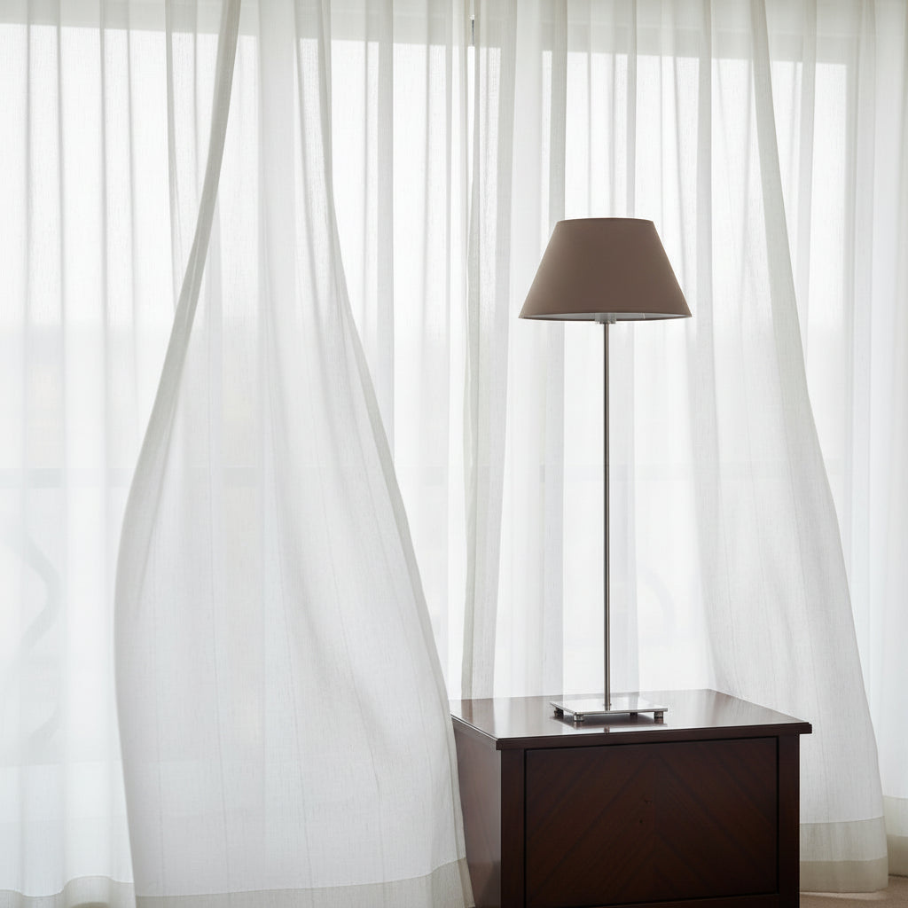 Table lamp with a brown shade sitting on dark bedside cabinet with white billowing drapes