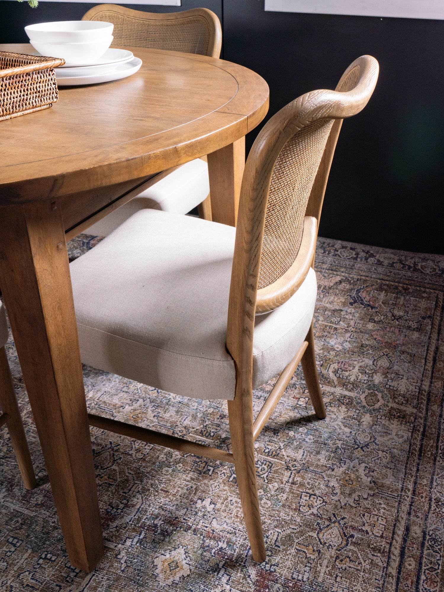 Wooden dining table with a chair featuring a wicker backrest on a patterned rug.