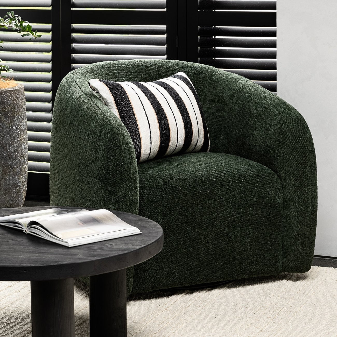 Green armchair with a striped cushion next to a round dark wood side table in a room with white walls and black shutters.