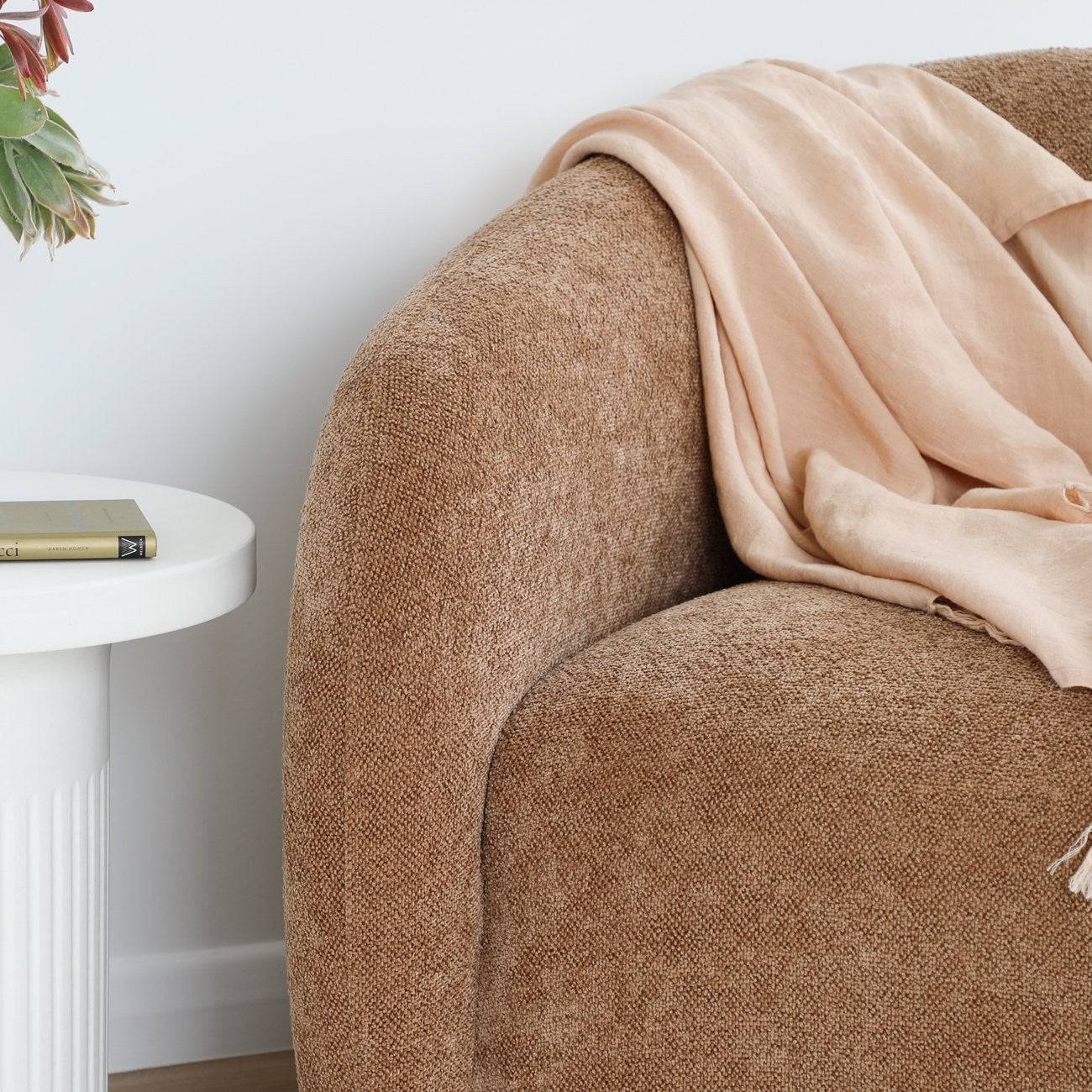 Caramel textured armchair with a beige throw draped over it, next to a white side table with a book.