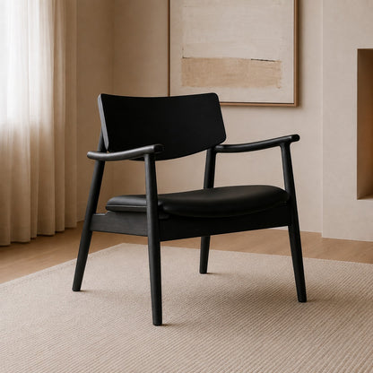 A modern black armchair with a curved wooden armrest and a low backrest, positioned on a textured beige rug in a minimalist room with warm beige walls and curtains.
