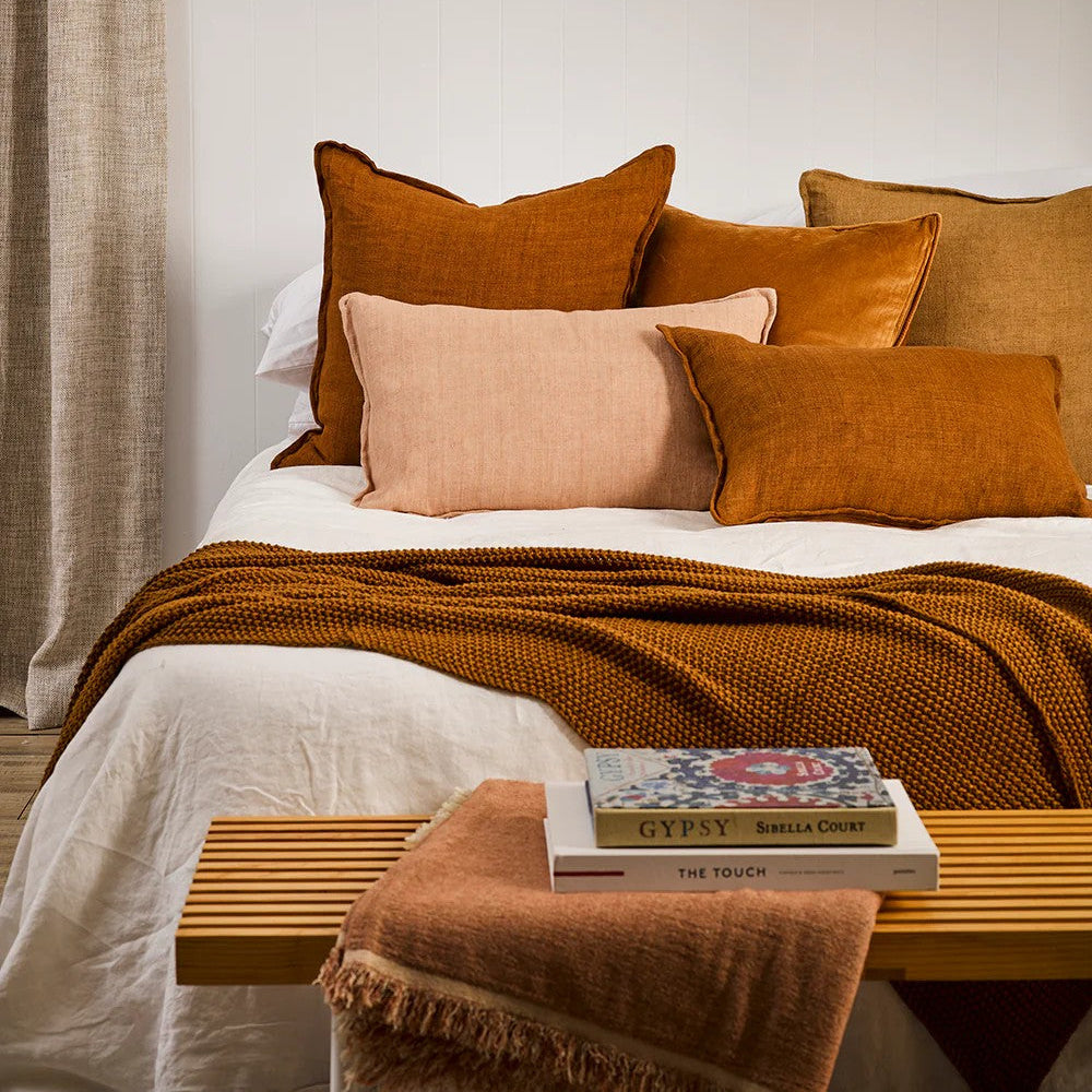 Bedroom with a bed featuring tobacco and salmon cushions and a blanket, next to a wooden bench with a blanket draped over it.