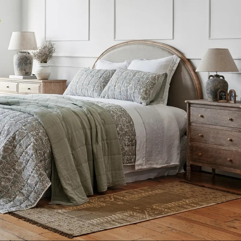 Bedroom with a bed featuring a green and grey patterned quilt, a curved top headboard, two bedside cabinets  with lamps, and a wooden floor.