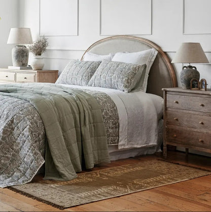 Bedroom with a bed featuring a green and grey patterned quilt, a curved top headboard, two bedside cabinets  with lamps, and a wooden floor.