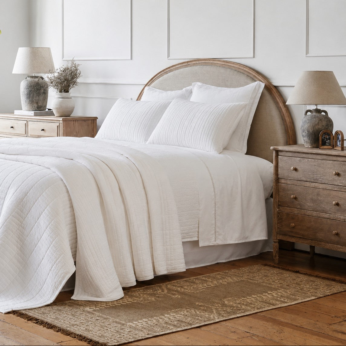 A neatly made bed dressed in white textured bedding and a white quilt, set in a softly styled bedroom with a linen and wood curved headboard, wooden bedside cabinets, lamps, panelled walls, and a patterned scatter rug.