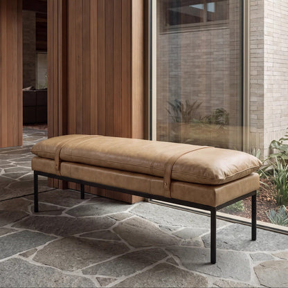 A modern tan leather bench seat with a black metal frame, positioned on a stone patio in front of a large window and wood-panelled wall.