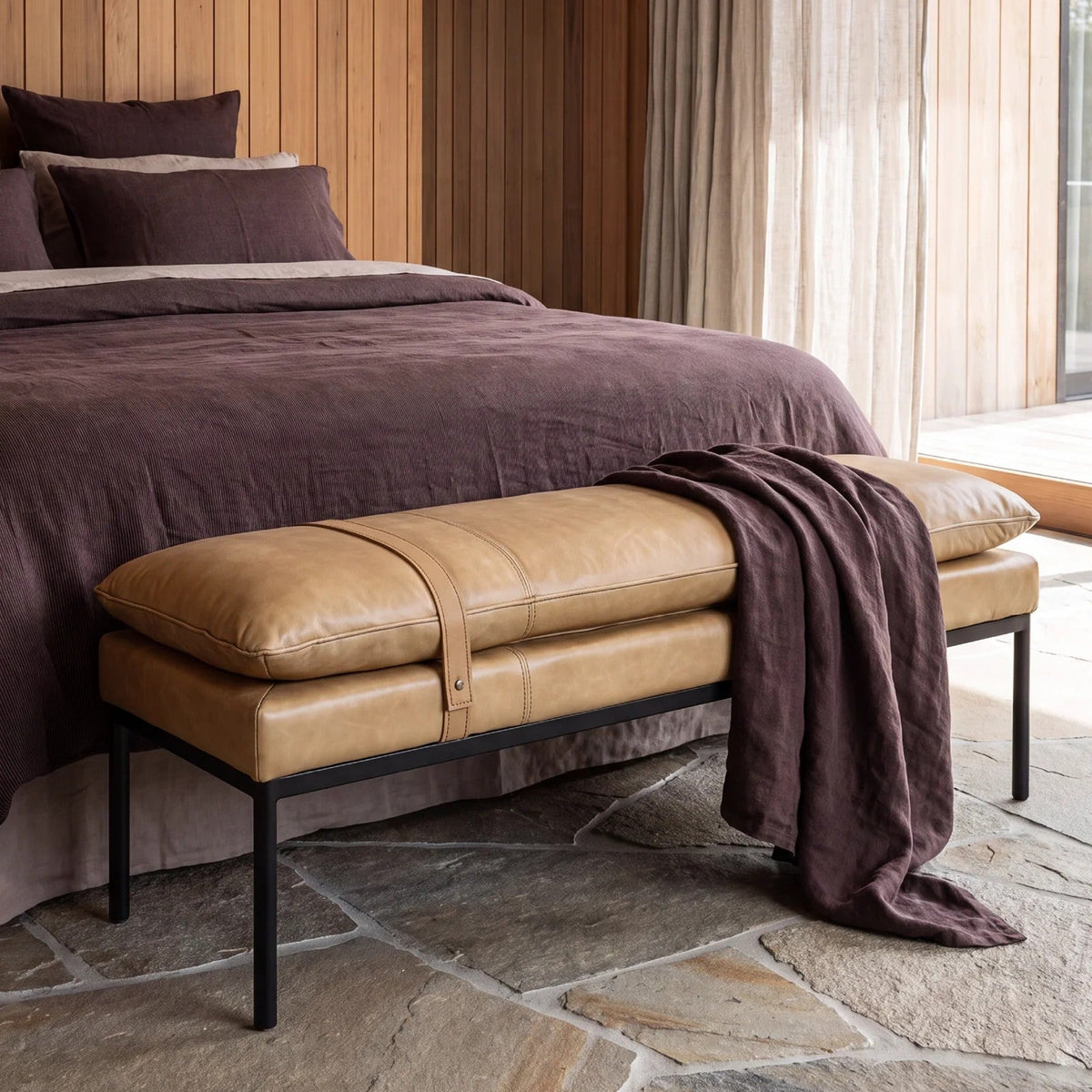 A tan leather bench with black metal legs sits at the foot of a bed in a rustic style bedroom with wood-panelled walls and stone flooring.