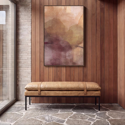 A framed abstract landscape painting with soft beige, brown, and muted purple tones hangs on a wood-panelled wall above a tan leather bench seat in a modern interior setting.