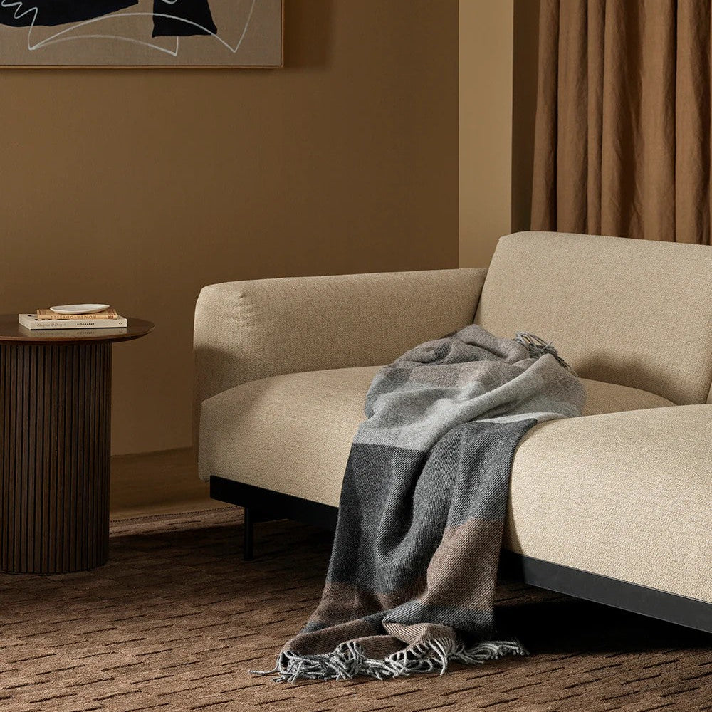 Beige sofa with a brown and grey plaid blanket draped over it, in a room with abstract art and a curtain.