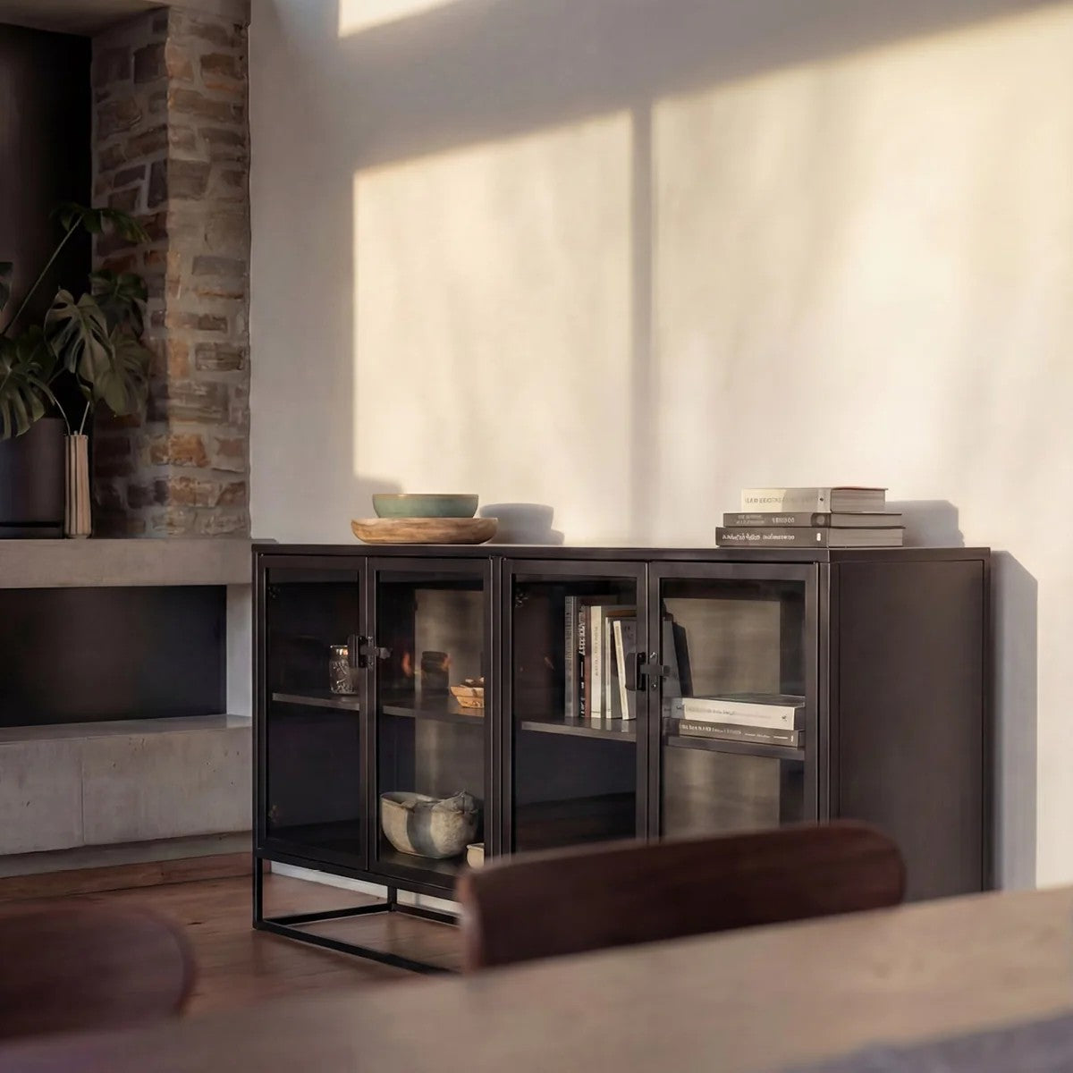 A black metal and glass sideboard styled in a modern living room setting with warm sunlight casting shadows on the wall.