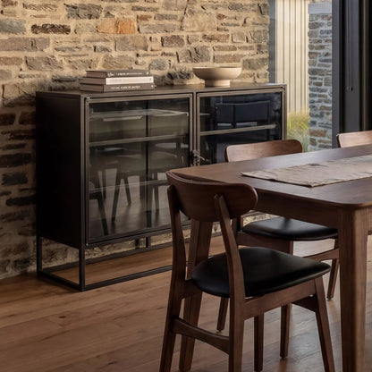 A dark wood dining room scene with a black metal and glass sideboard cabinet against a stone brick wall, next to a wooden dining table and chairs.