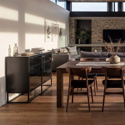 A modern dining room scene with a dark wood dining table and chairs, a black glass sideboard, and a gray sofa in a sunlit interior with wood floors and a stone accent wall.