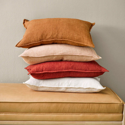 Stack of four pillows in almond, red, coconut and tobacco e on a tan bench against a beige wall.