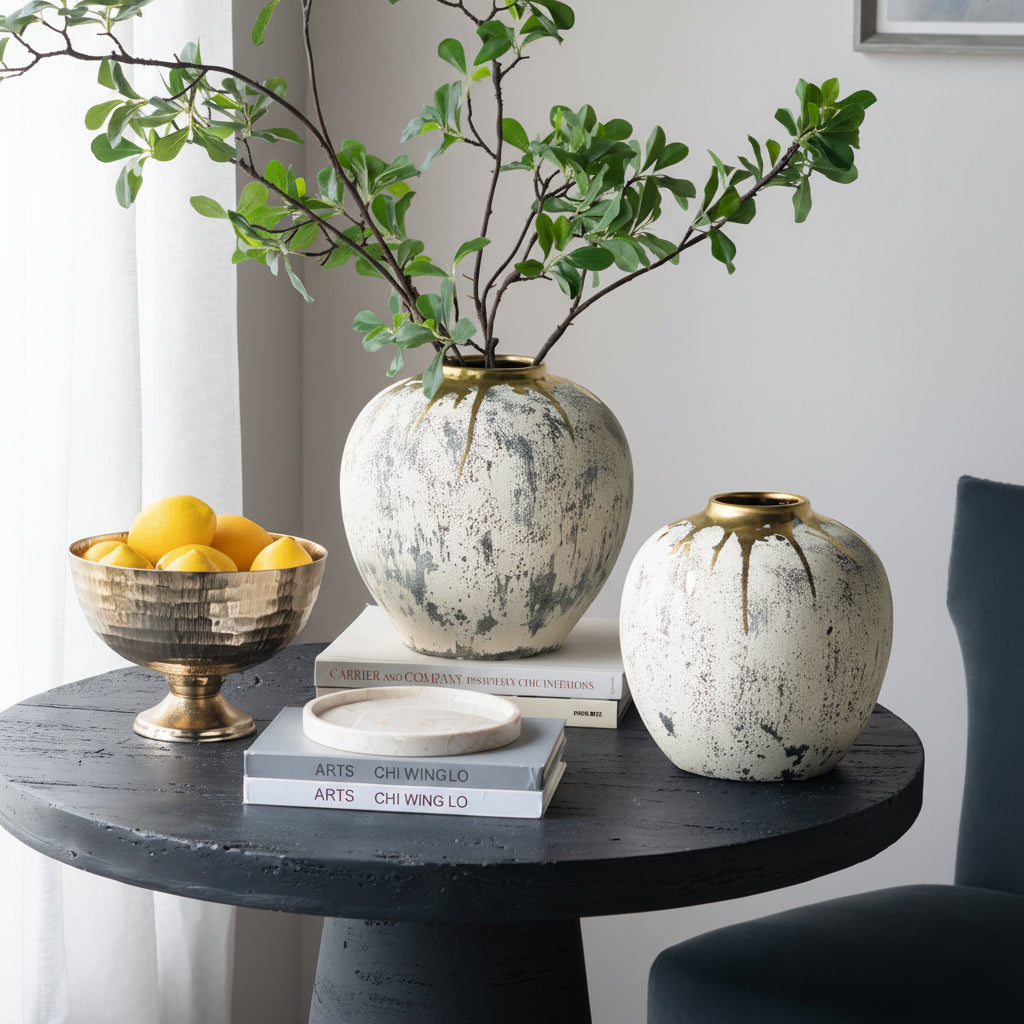 Decorative table setting with ceramic vases, a bowl of fruit, and books against a neutral wall.