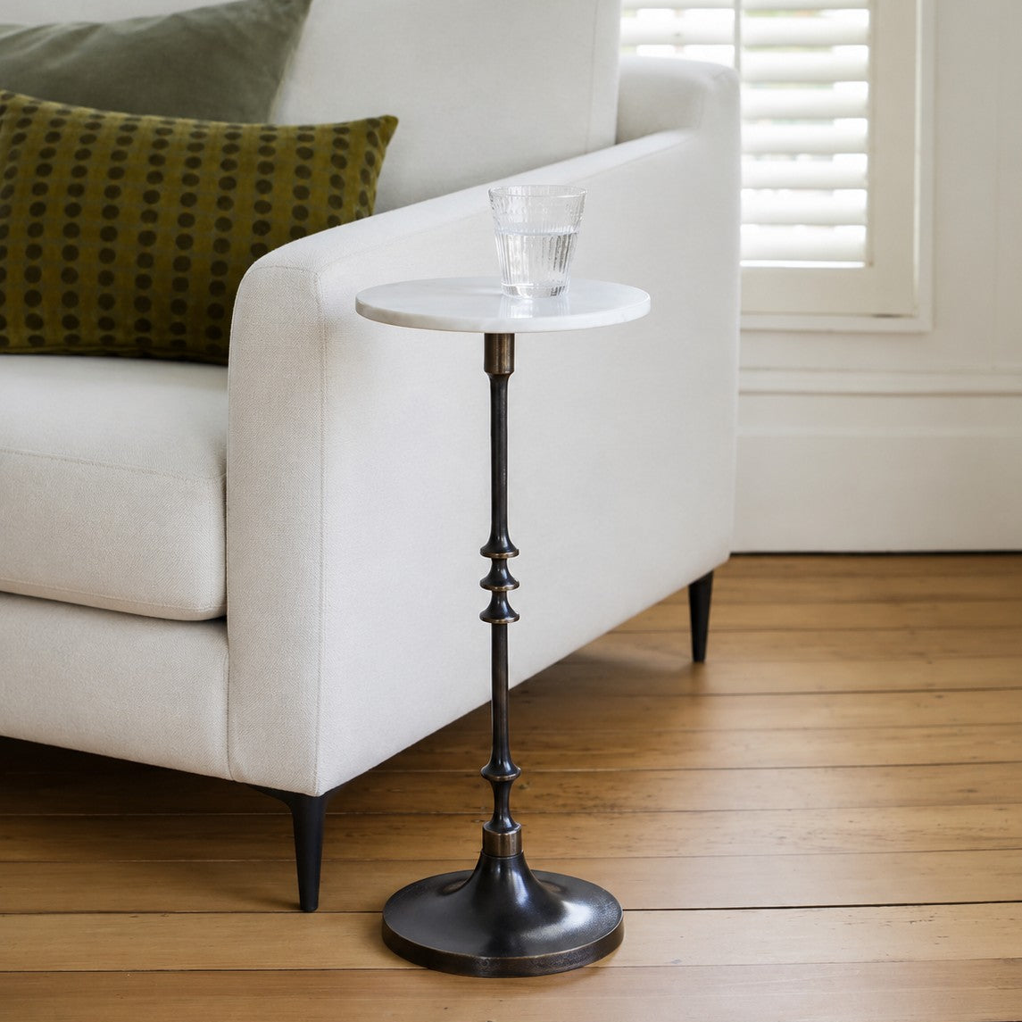 A small round side table with a white marble top and a dark metal pedestal base, styled next to a white armchair on a wooden floor.
