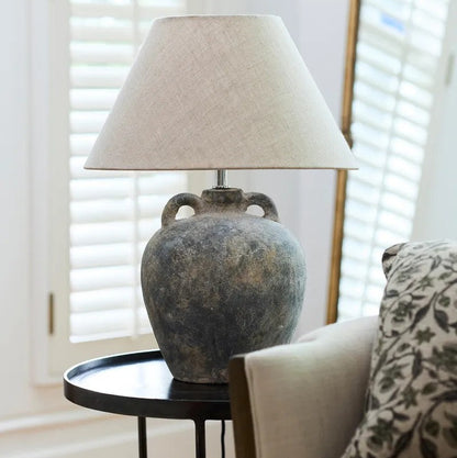 Table lamp with a textured base on a small round table in a room with light-coloured walls and a window with shutters.