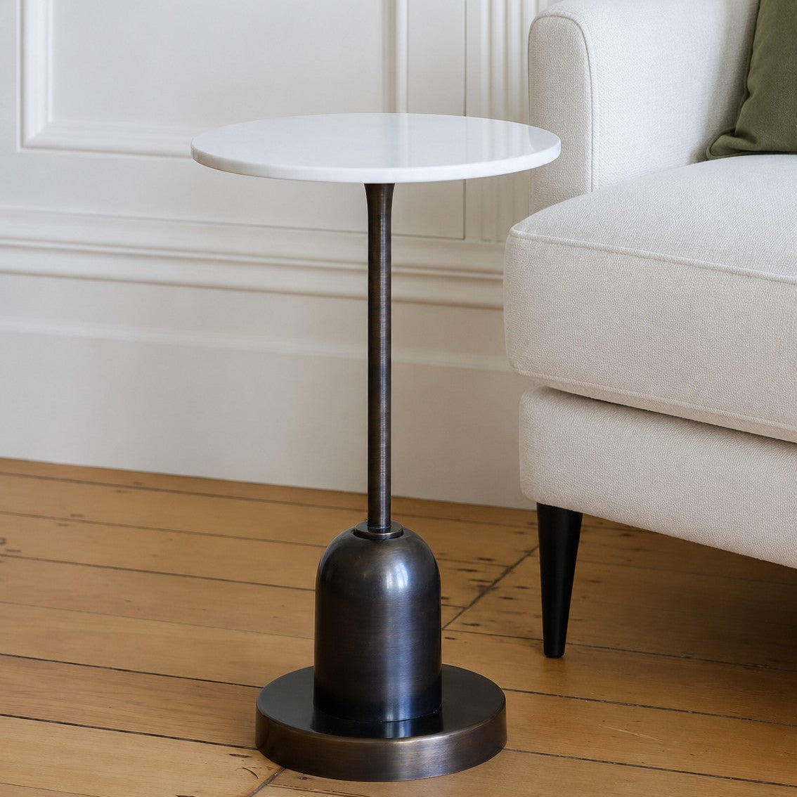 A small round side table with a white marble top and a dark bronze pedestal base, shown in a styled living room next to a light-colored armchair on a wooden floor.