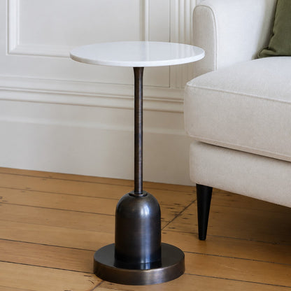 A small round side table with a white marble top and a dark bronze pedestal base, shown in a styled living room next to a light-colored armchair on a wooden floor.
