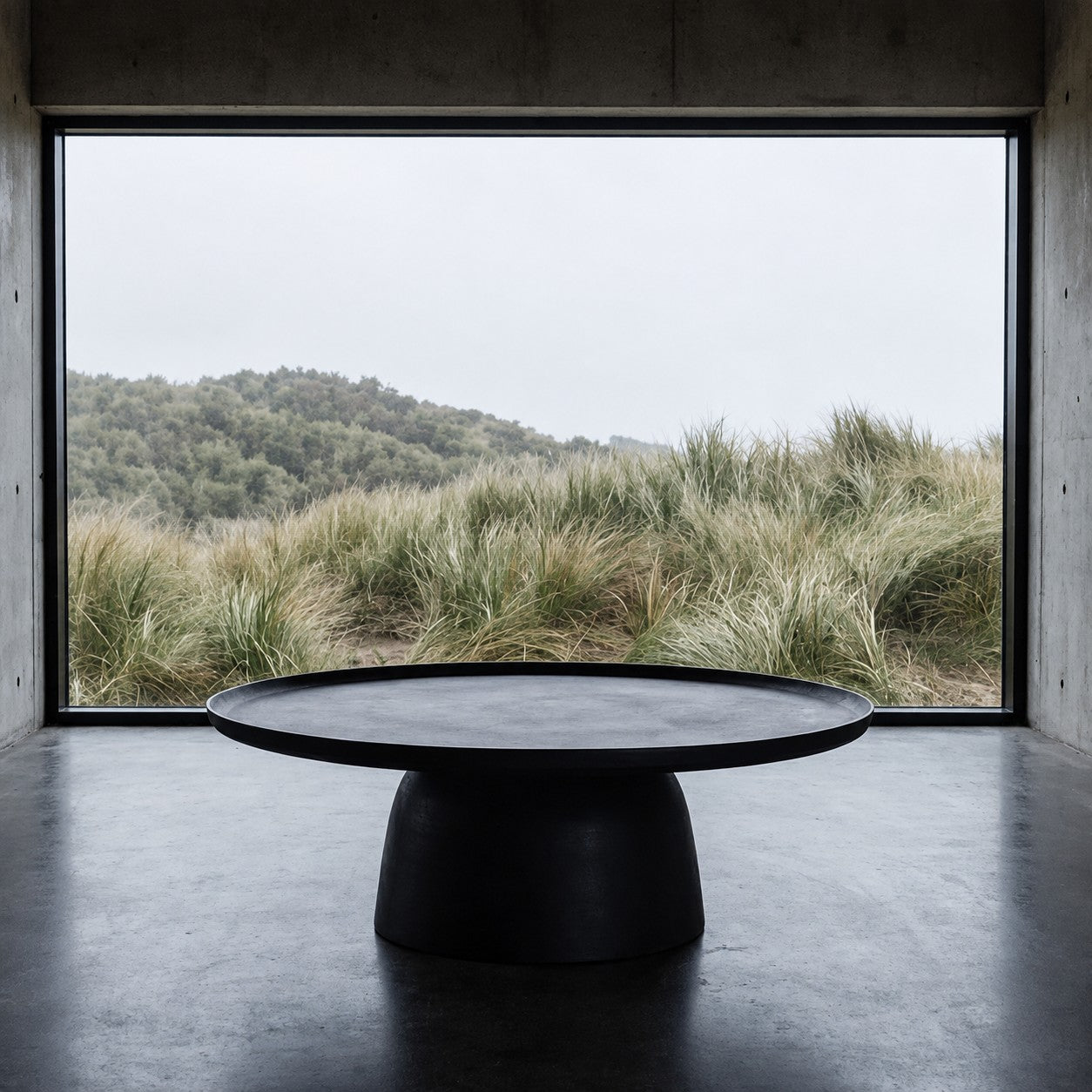 A round black aluminium coffee table sits in a minimalist concrete room, looking out through a large window to a sand dune landscape under an overcast sky.