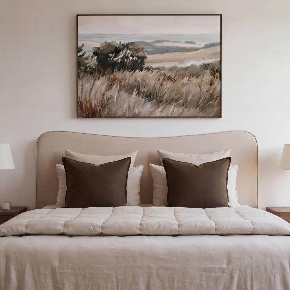 A framed landscape painting of a grassy field hangs above a bed with a beige upholstered headboard, cream bedding, and two dark brown pillows in a softly lit bedroom.