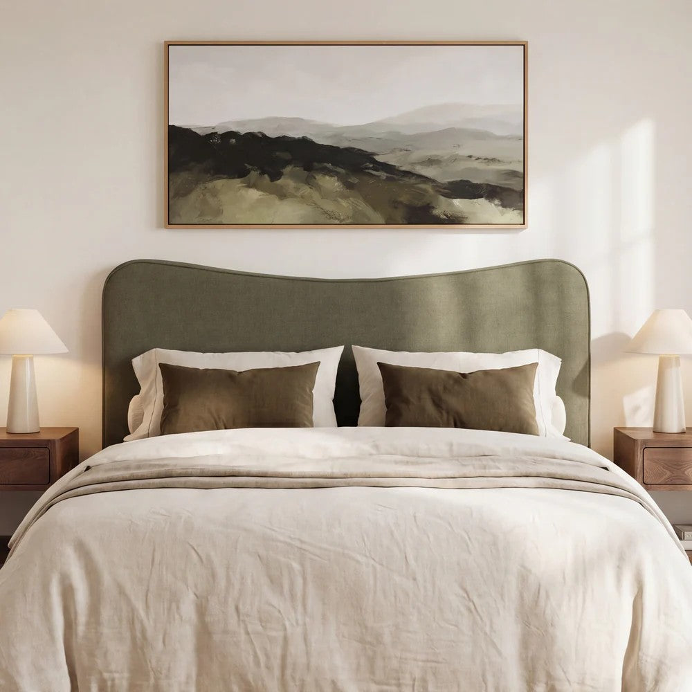 A styled bedroom scene featuring a large framed landscape painting above a bed with a muted herb green headboard, cream bedding, and matching pillows.