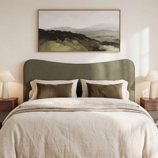 A styled bedroom scene featuring a large framed landscape painting above a bed with a muted herb green headboard, cream bedding, and matching pillows.
