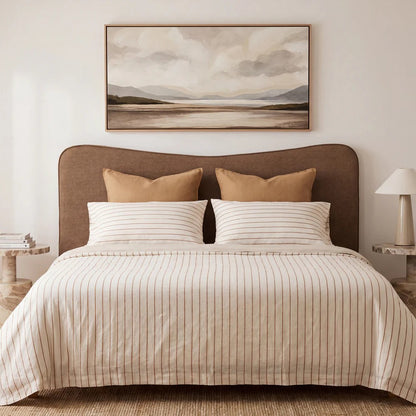 A neatly made bed with a cream and tan striped duvet cover and matching pillows, set in a minimalist bedroom with a toffee brown upholstered headboard, a framed landscape painting above, and bedside tables with lamps.
