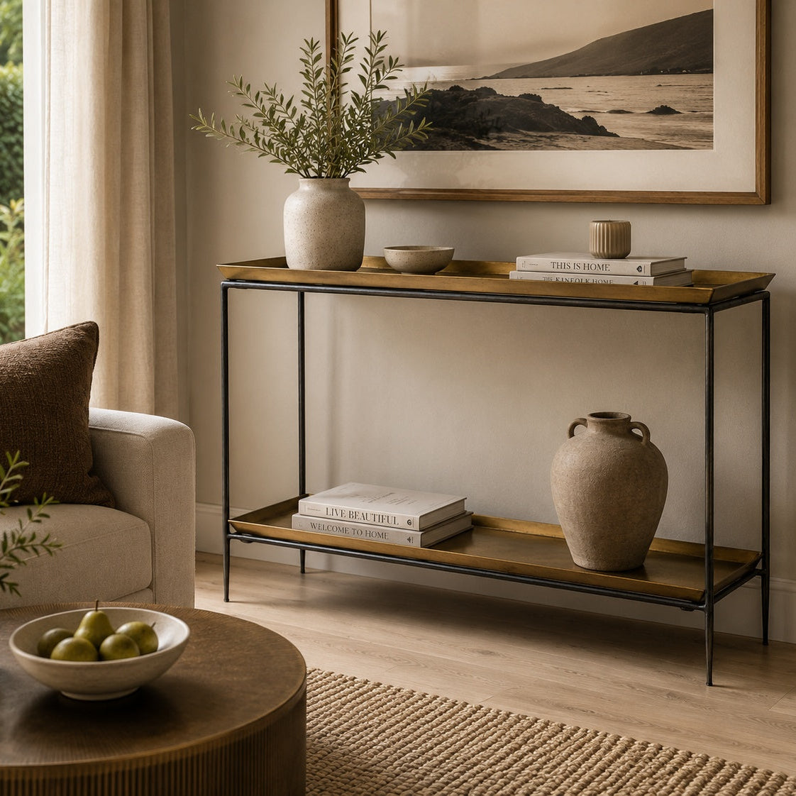 A two-tier console table with a black metal frame and gold painted wood shelves, styled in a warm, modern living room setting with home decor and books.