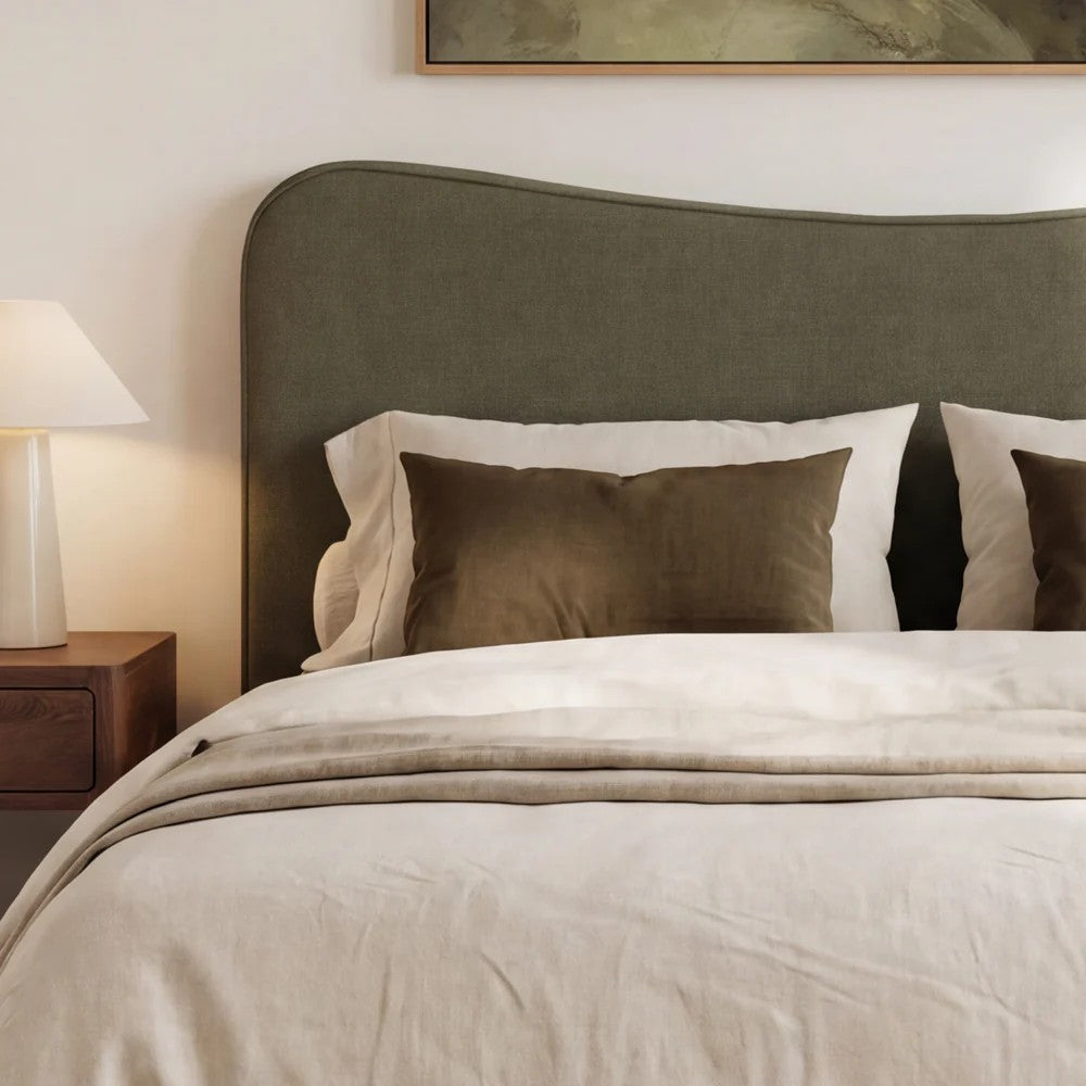 A styled bedroom scene showing a bed with a herb coloured upholstered  headboard, cream bedding, two dark brown pillows, a bedside lamp, and part of framed wall art above.