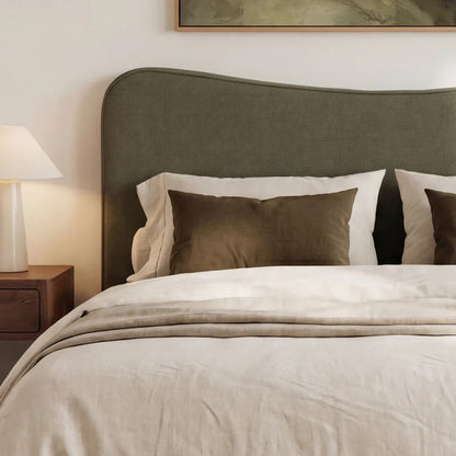 A styled bedroom scene showing a bed with a herb coloured upholstered  headboard, cream bedding, two dark brown pillows, a bedside lamp, and part of framed wall art above.