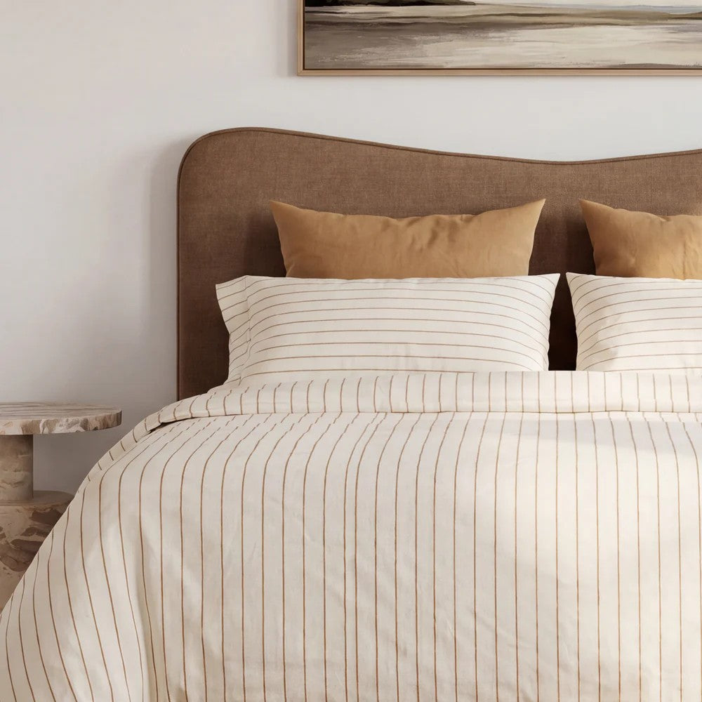 A neatly made bed in a bedroom setting, featuring a cream-colored duvet cover and matching pillowcases with thin vertical brown stripes, styled with two tan accent pillows against a toffee brown upholstered headboard.