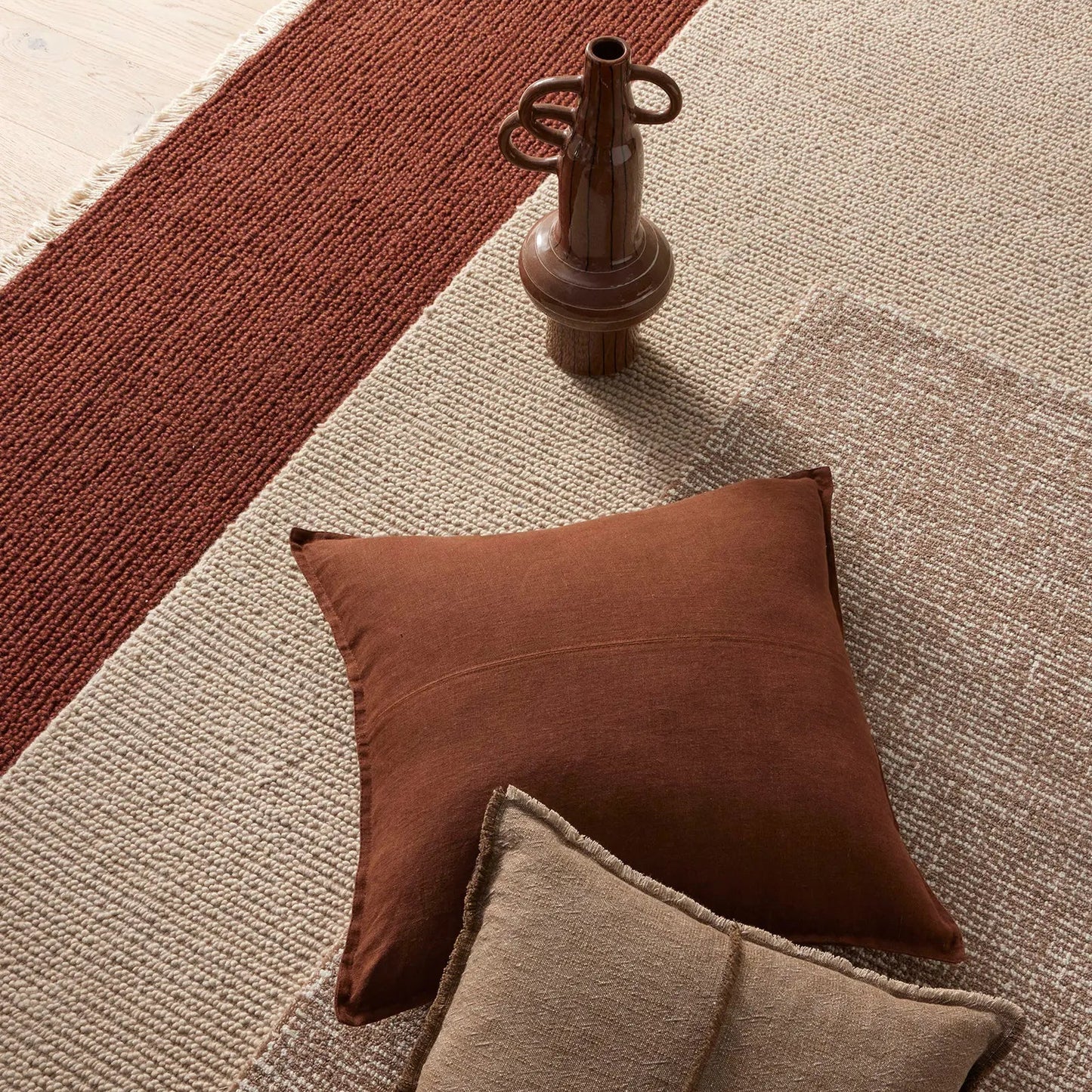 Brown vase on a patterned rug with two cushions.
