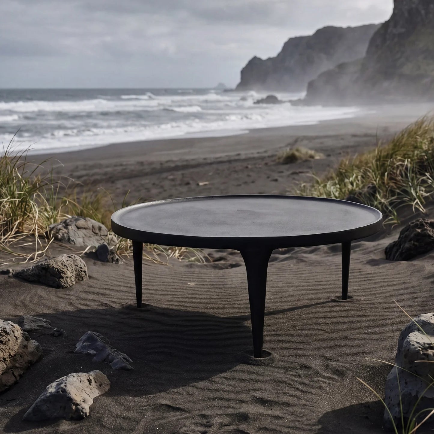 A round black outdoor coffee table with three legs sits on a sandy beach with ocean waves and rocky cliffs in the background.