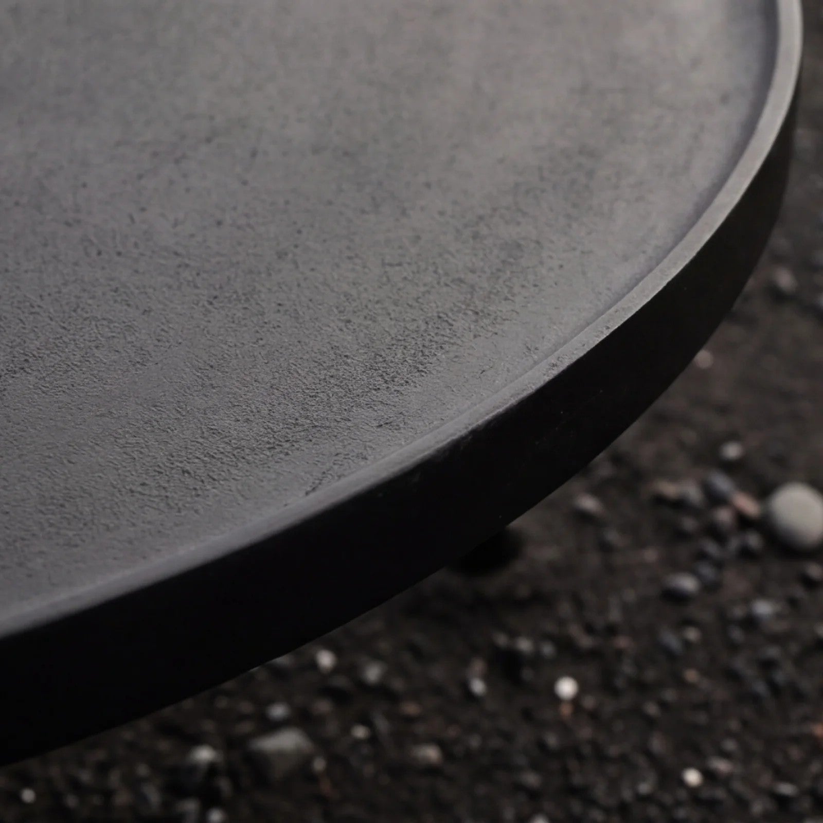Close-up of a round black aluminium coffee table with a textured matte finish, shown on a dark gravel surface.