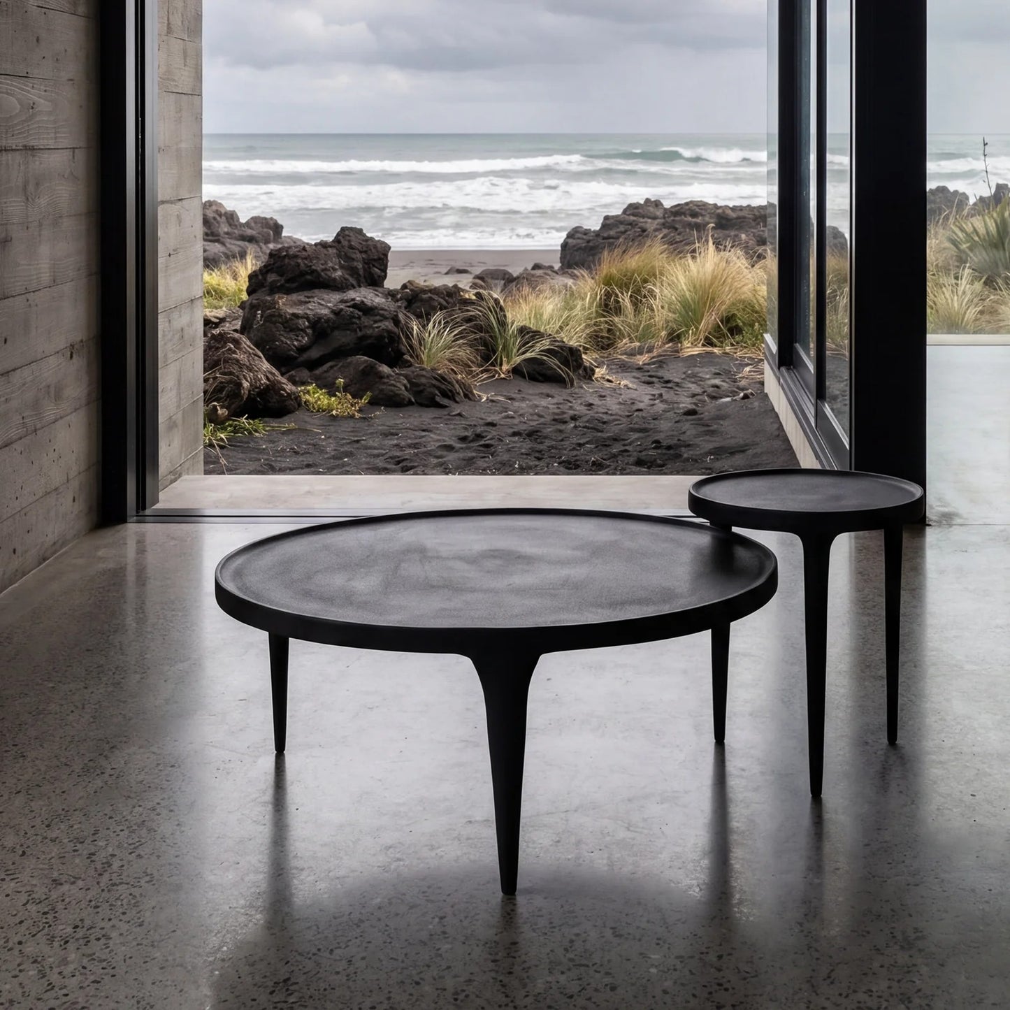 Two round black nesting coffee tables with tapered legs, set inside a modern concrete room with a large window overlooking a rocky beach and ocean.