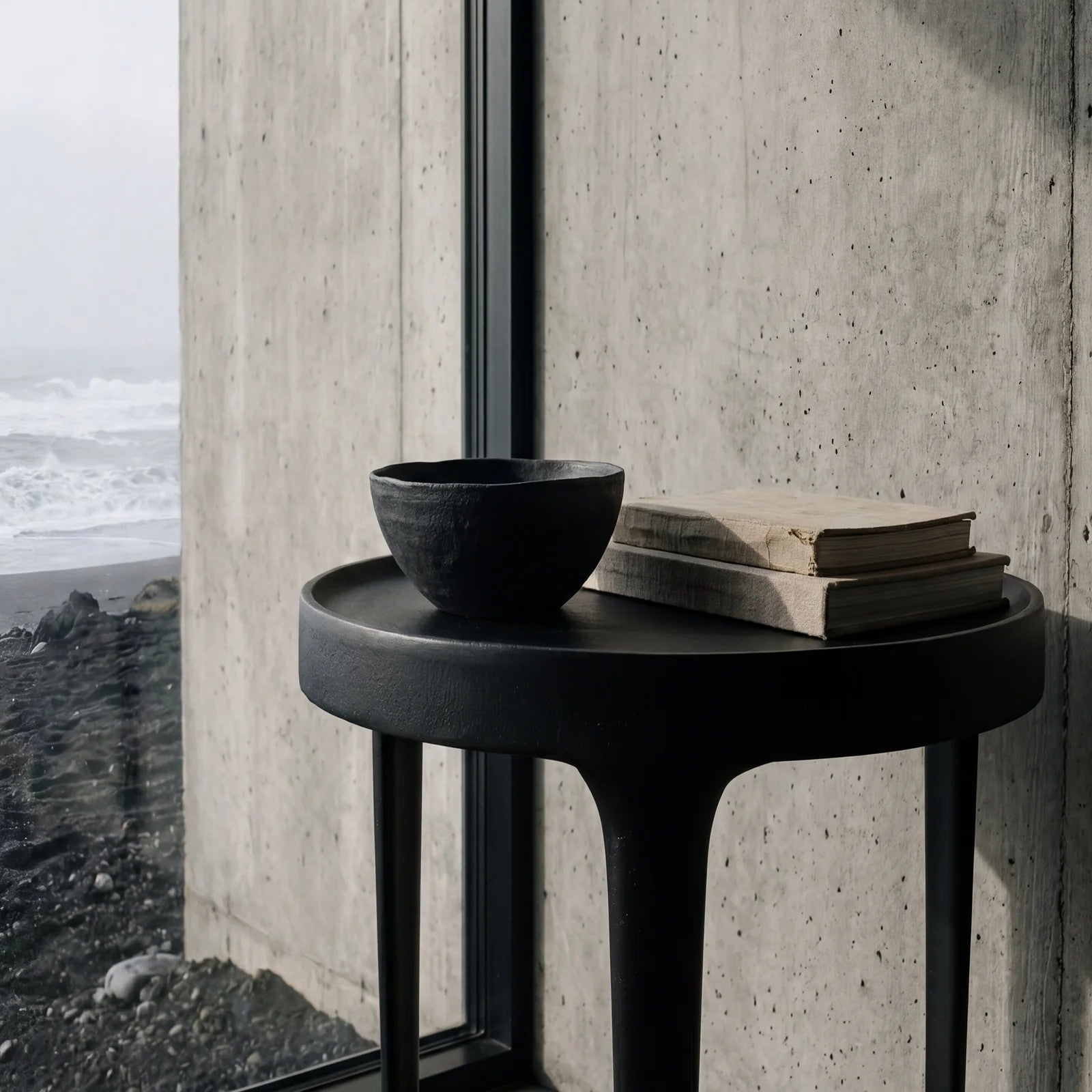 A round black side table with a matte finish, styled with a black ceramic bowl and two stacked vintage books, positioned next to a large window showing a black sand beach and ocean view.
