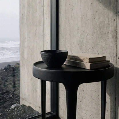 A round black side table with a matte finish, styled with a black ceramic bowl and two stacked vintage books, positioned next to a large window showing a black sand beach and ocean view.