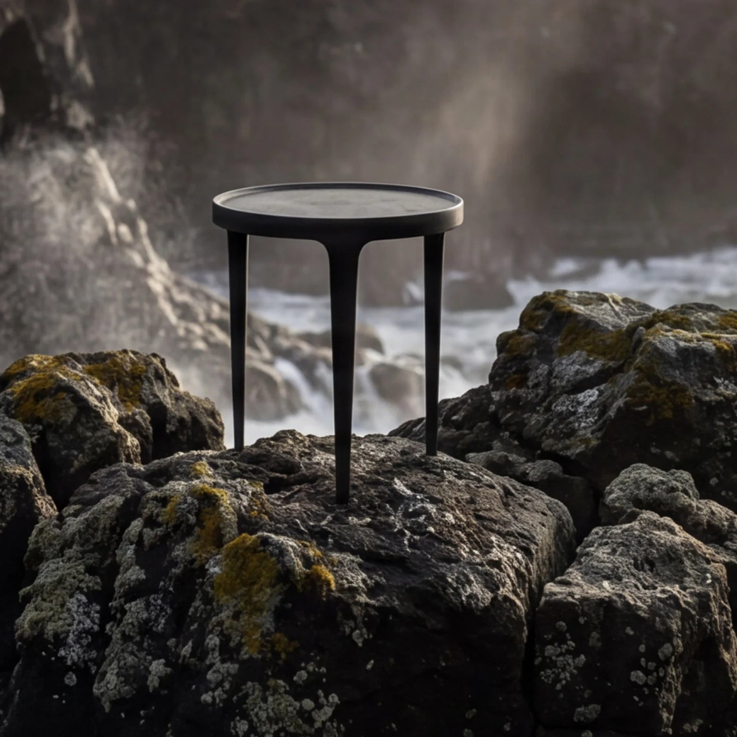 A round black metal side table with three legs stands on dark coastal rocks with a misty ocean scene in the background.