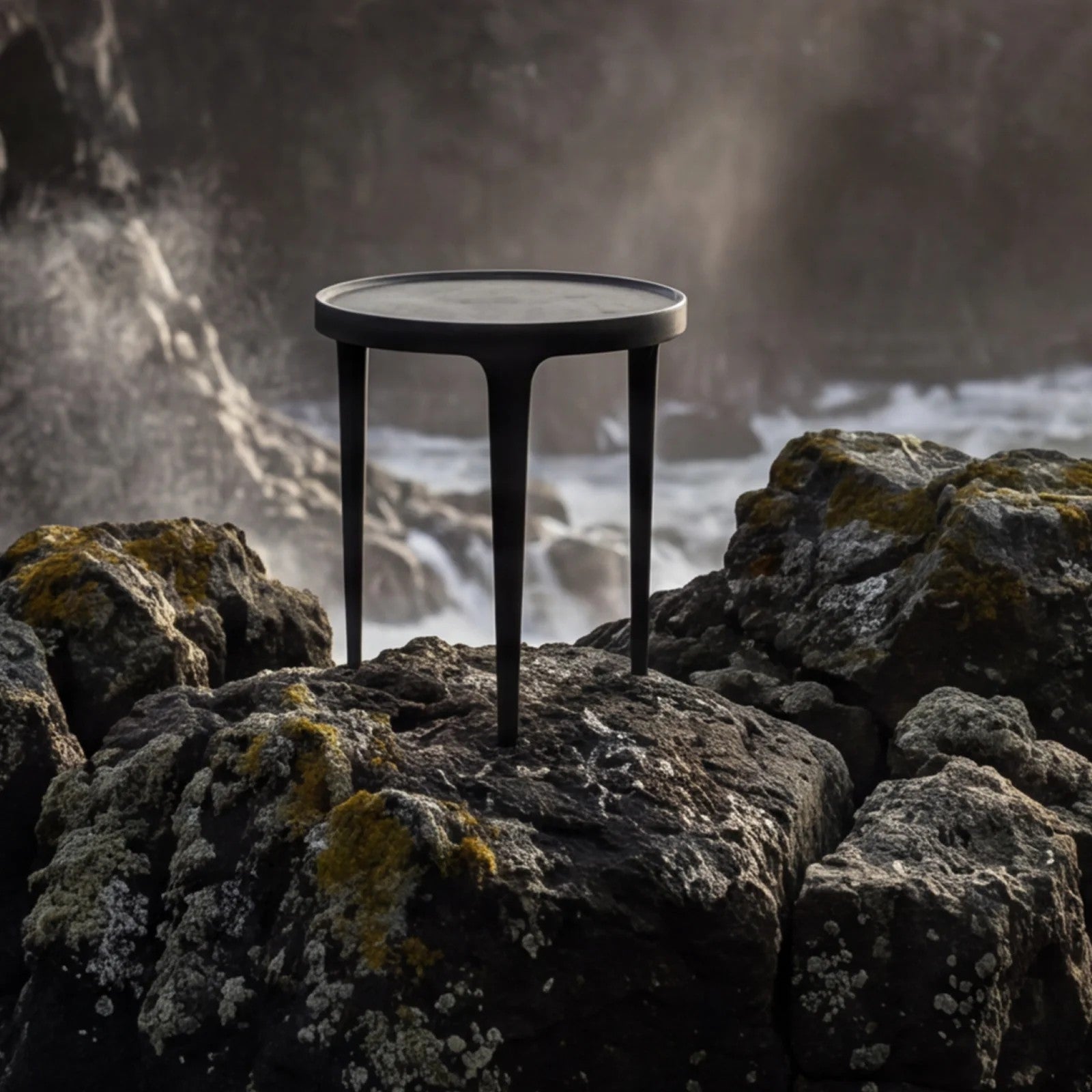 A round black metal side table with three legs stands on dark coastal rocks with a misty ocean scene in the background.