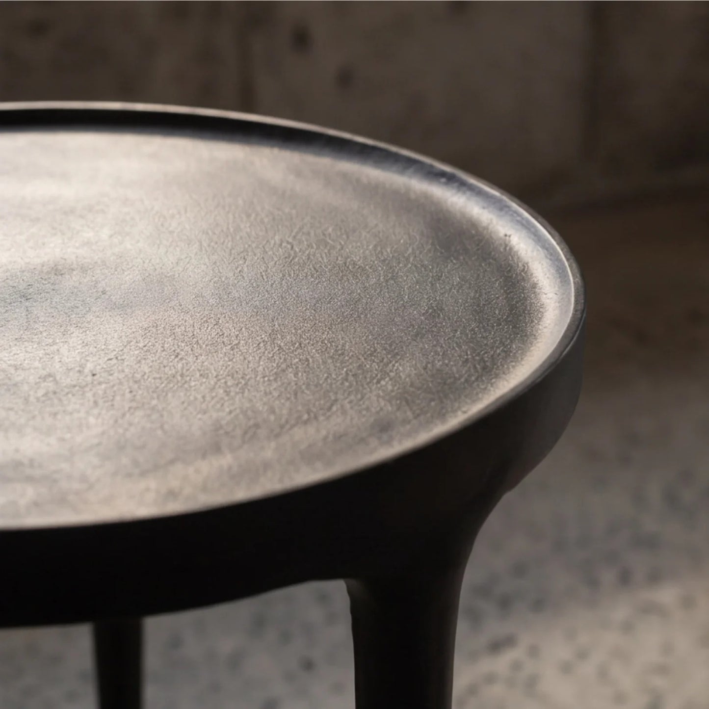 Close-up of a black side table top with a textured matte finish and a thick rim, supported by dark legs, set against a blurred concrete background.